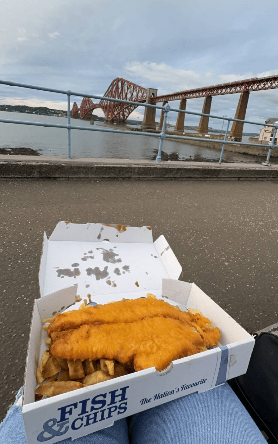 A box of Maurizio’s SQ fish and chips with the Forth Bridge visible in the background over water.